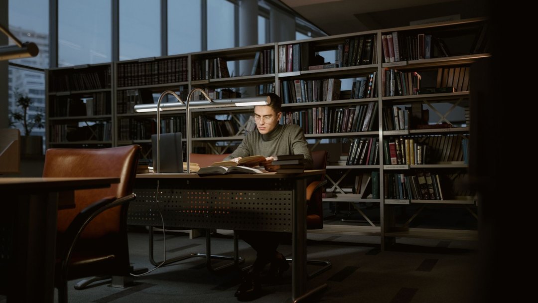 图 / Pexels · Tima Miroshnichenko A man intensely studies in a quiet library, surrounded by books and soft lighting.