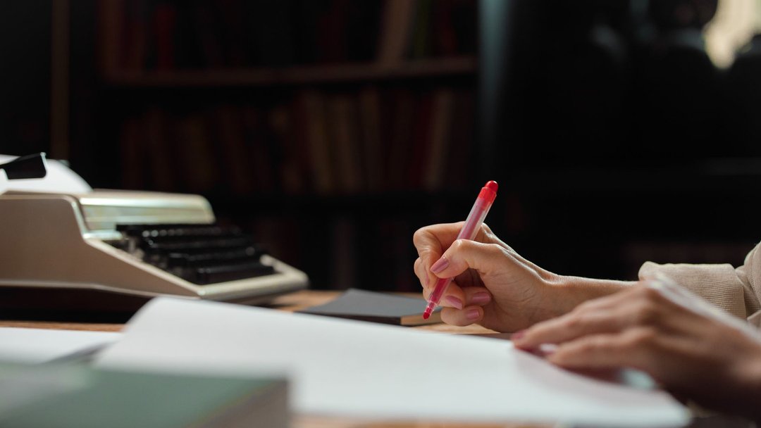 图 / Pexels · Ron Lach A hand writing with a pen in a cozy library setting with a vintage typewriter.