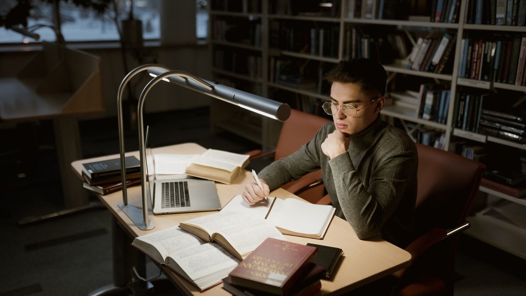 图 / Pexels · Tima Miroshnichenko A young adult studying at a library desk with books and laptop under warm lighting.