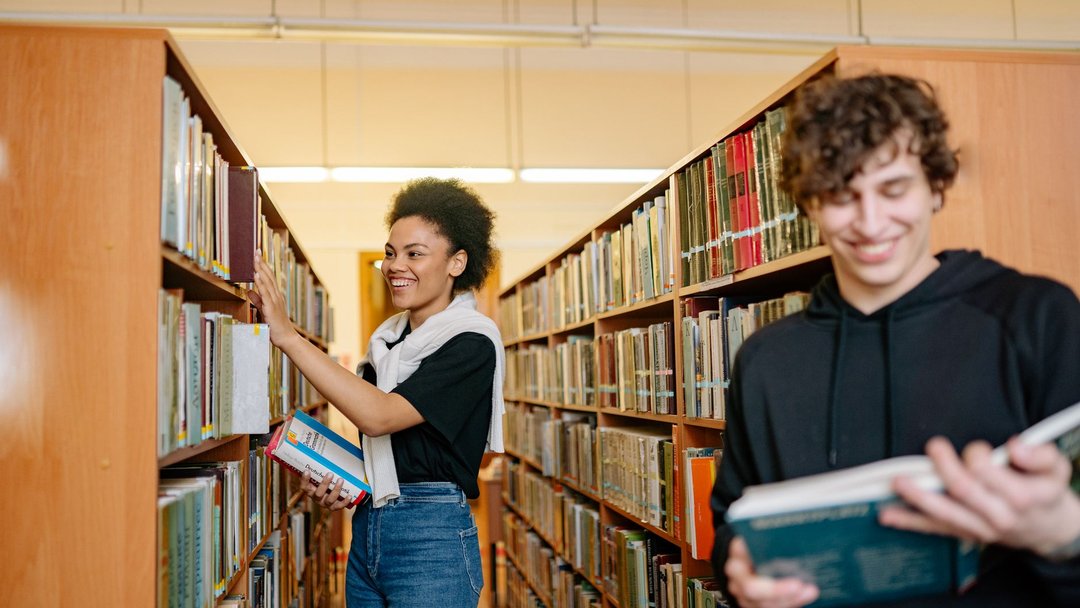 图 / Pexels · Yan Krukau Two students studying in a university library, choosing books from shelves.