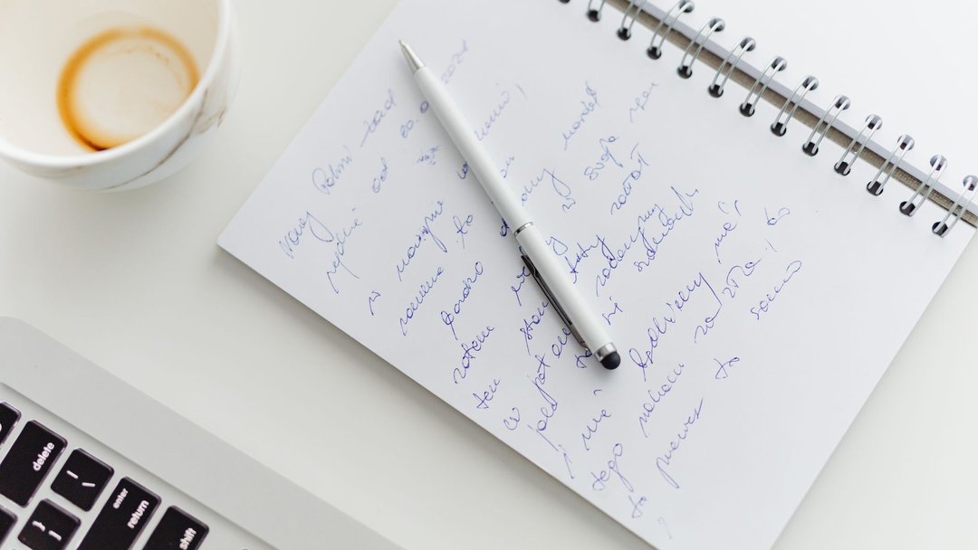 Close up of a notebook with handwriting, a pen, coffee cup, and laptop on a white desk.