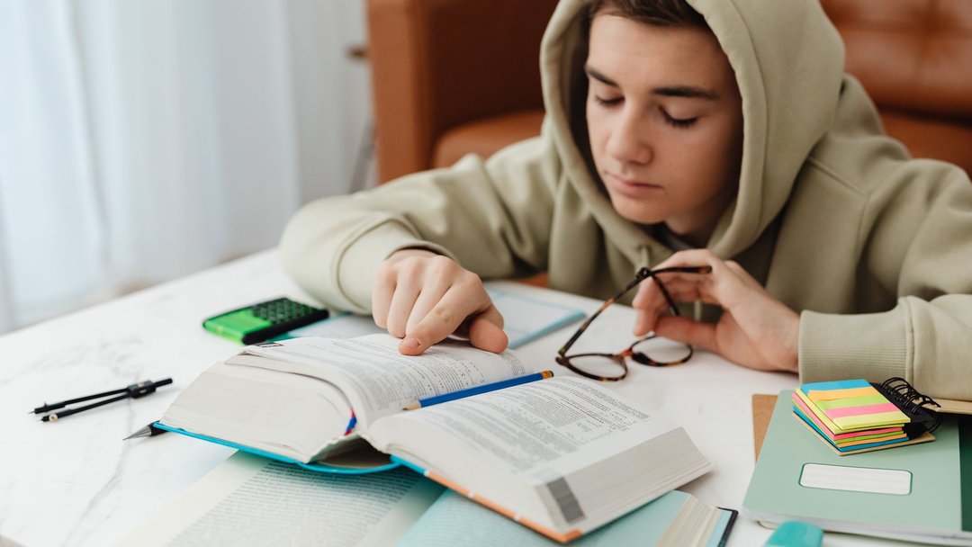 图 / Pexels · https://kaboompics.com/ A teenager in a hoodie studying with books and notes on a table, focused on learning indoors.